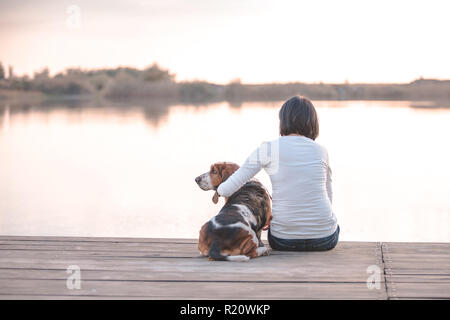 Piuttosto giovane donna che abbraccia il suo cane Basset Hound sul dock di legno sul fiume Foto Stock