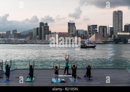 Hong Kong, Cina - 16 Maggio 2018: persone facendo Yoga di gruppo in un parco che si affaccia sul porto Victoria con lo skyline di Kowloon in Isola di Hong Kong Foto Stock