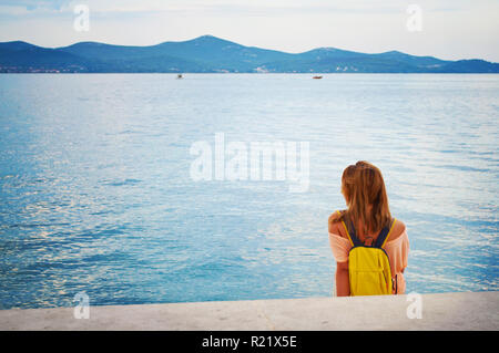 Una ragazza giovane con uno zaino seduta pacificamente su scalini in pietra vicino la riva del mare guardando wistfully presso la collina di gamma e yacht in distanc Foto Stock