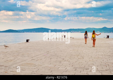 Due ragazze camminare tra i gabbiani battenti vicino la riva del mare sullo sfondo di una collina di gamma e drammatico mattino cielo nuvoloso. Zadar, Croazia Foto Stock