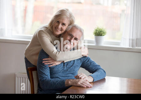Ritratto di senior sorridente abbracciando la moglie amorevole marito da dietro a casa di età felice coppia abbracciare guardando la telecamera esprimendo il sostegno e la cura, e Foto Stock