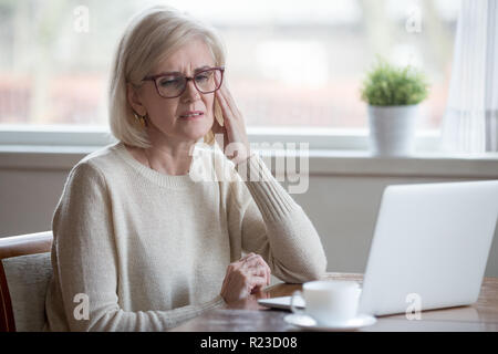Sconvolto di età della donna utilizzando portatile a casa che soffrono di mal di testa, stanchi senior femminili templi di massaggio avente forti dolori o visione offuscata, esausto el Foto Stock