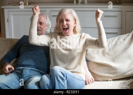 Felice di età matura rilassarvi sul lettino accogliente a casa a guardare partita di calcio in TV, eccitato senior moglie urlare il tifo squadra preferita, anziane donne s Foto Stock