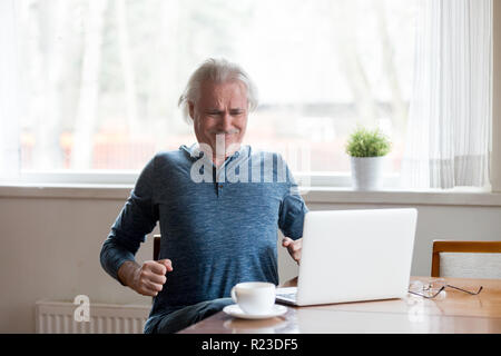 Esaurito uomo più anziano che soffrono di dolori alla schiena seduta troppo a lungo nella postura non corretta, stanchi senior tratto maschio in sedia a lavorare al computer portatile avente corpo sp Foto Stock