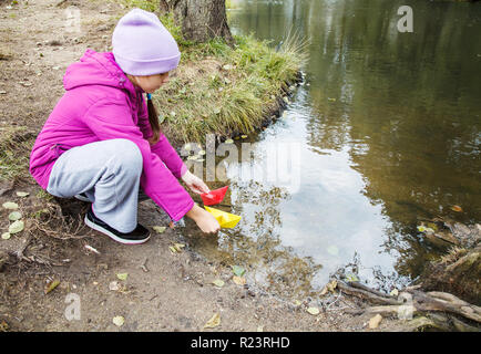 Ragazza in blu hat, magenta giacca e pantaloni grigi messa due colorate barche di carta nel flusso all'esterno sul giorno di autunno Foto Stock