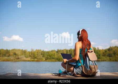 Immagine sul retro della donna nelle cuffie con zaino e laptop in mani seduto sulla riva del fiume Foto Stock
