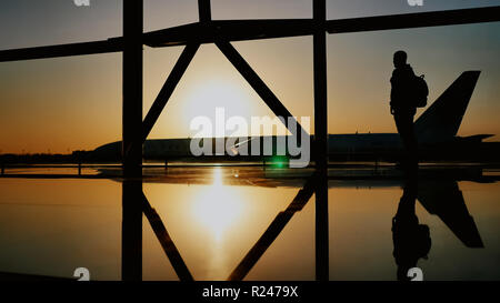 La silhouette del turista guy guardando il decollo del piano e passando un enorme piano in piedi alla finestra di airport al tramonto di sera. Il concetto di viaggio, la gente in aeroporto. Foto Stock