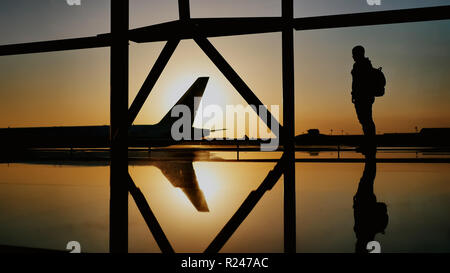 La silhouette del turista guy guardando il decollo del piano e passando un enorme piano in piedi alla finestra di airport al tramonto di sera. Il concetto di viaggio, la gente in aeroporto. Foto Stock