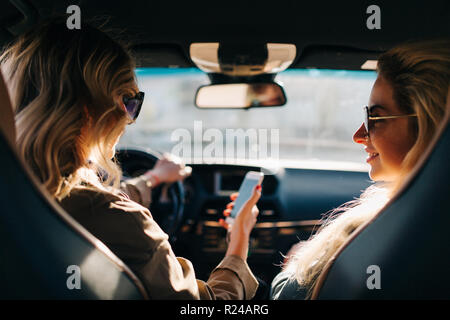 Foto della parte posteriore di due donne con capelli lunghi e il telefono in mano seduti in auto al sedile anteriore Foto Stock