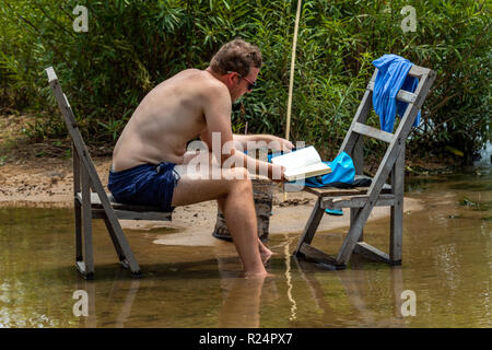 Don Det, Laos - Aprile 25, 2018: Turistica seduti su sedie di legno su una banca del fiume Mekong e la lettura di un libro Foto Stock