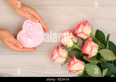 Donna di mani tenendo una confezione regalo a forma di cuore su grigio Sfondo di legno con bellissime rose. Concetto di dare un regalo per le vacanze. Vista dall'alto. Foto Stock