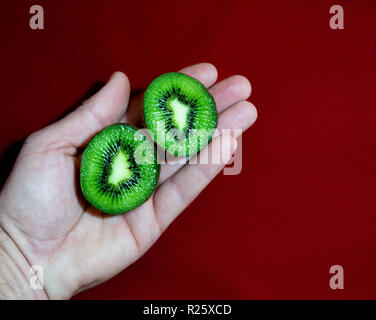 Primo piano di una mano con due metà tagliare kiwi su uno sfondo rosso Foto Stock