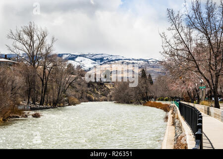 Truckee fiume che scorre attraverso il centro cittadino di Reno su un nuvoloso giorno di primavera, Nevada; lastricato sentiero sulla destra; Sierra montagne coperte di neve in Foto Stock