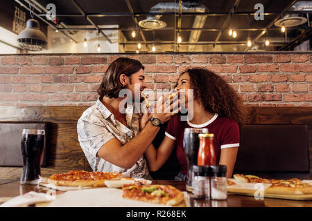 Giovane uomo e donna seduta presso un ristorante fast food per alimentare ogni altro con la pizza. Giovane godendo di mangiare la pizza al cafe. Foto Stock