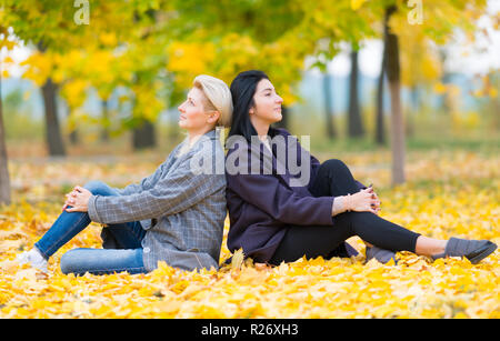 Due giovani donne seduta di schiena in posizione di parcheggio sul suolo coperto di colore giallo brillante le foglie in autunno, visto dal lato Foto Stock