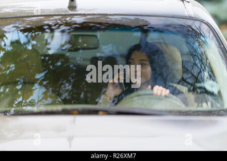 Giovane donna conducente chat sul suo telefono cellulare mentre si guida visto dalla parte anteriore attraverso il parabrezza Foto Stock