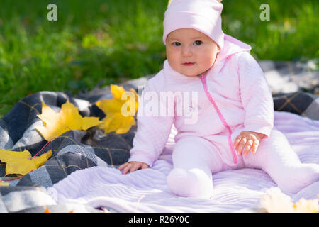 Curioso little Baby girl fissando la telecamera come si siede su una coperta all'aperto in giardino durante l'autunno in un grazioso abito rosa Foto Stock