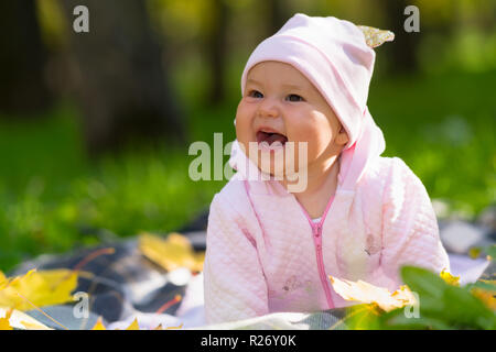 Laughing baby girl con un ampio sorriso raggiante giocando su una coperta sul prato di un autunno nel parco un ritratti Foto Stock