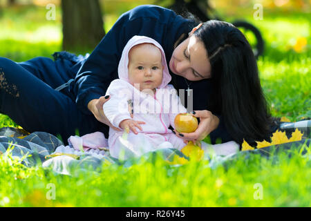 Giovane madre con il suo neonato in un parco seduto su un tappeto in una lussureggiante verde erba offrire al bambino una Fresca mela autunnale Foto Stock