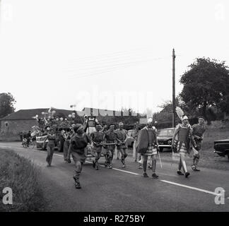 1967, storico villaggio inglese carnevale, persone vestite come centurioni romani a piedi lungo una strada di campagna che prendono parte ad un villaggio locale parade, Oxfordshire, Inghilterra, Regno Unito. Foto Stock