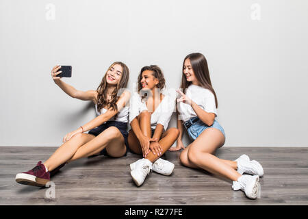 Immagine dei tre giovani carino belle ragazze amici seduti isolate su sfondo bianco rendono selfie dal telefono cellulare. Foto Stock