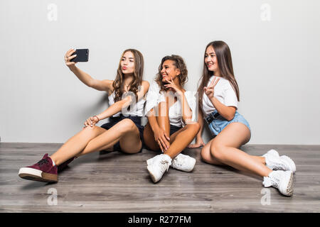 Immagine dei tre giovani carino belle ragazze amici seduti isolate su sfondo bianco rendono selfie dal telefono cellulare. Foto Stock