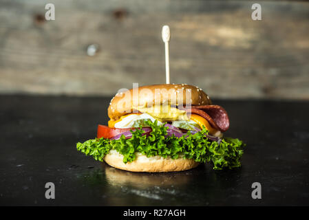 Gustosa cheesburger con un gran numero di verdure, senape, maionese, salame e viola per anelli di cipolla in legno scuro dello sfondo Foto Stock