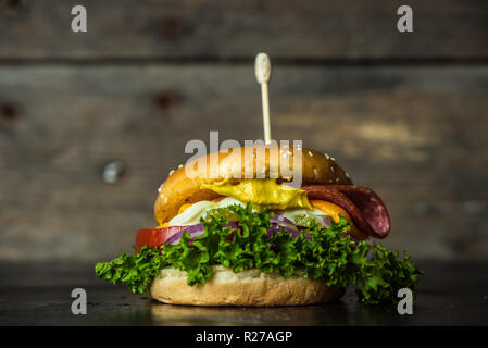 Gustosa cheesburger con un gran numero di verdure, senape, maionese, salame e viola per anelli di cipolla in legno scuro dello sfondo Foto Stock