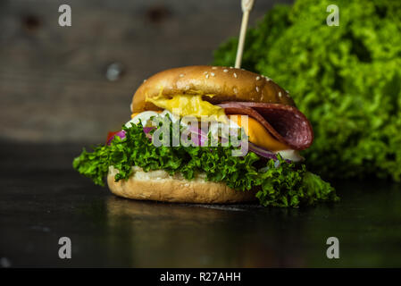 Gustosa cheesburger con un gran numero di verdure, senape, maionese, salame e viola per anelli di cipolla in legno scuro dello sfondo Foto Stock