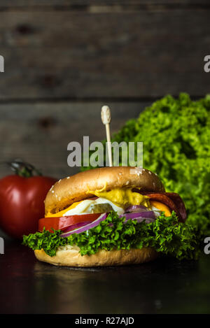 Gustosa cheesburger con un gran numero di verdure, senape, maionese, salame e viola per anelli di cipolla in legno scuro dello sfondo Foto Stock