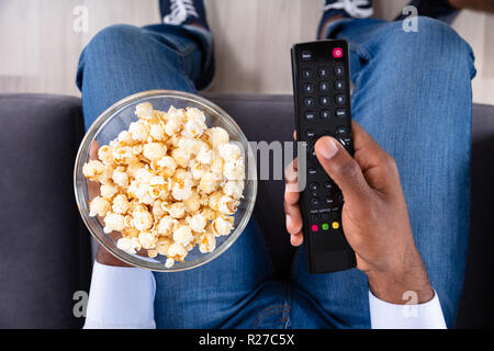 Alta Vista angolare di una mano di un uomo azienda telecomando della televisione e una ciotola di pop-corn Foto Stock