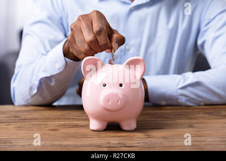 Mano d'uomo inserimento di moneta in Piggybank sulla scrivania in legno Foto Stock