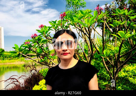 Una donna cinese in piedi di fronte ad una Plumeria rubra o rosso frangipani impianto entro Futian Mangrove Parco Ecologico di Shenzhen in Cina su un soleggiato blu Foto Stock