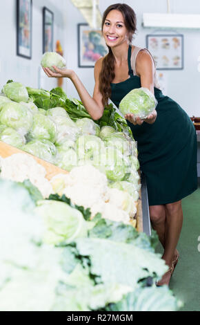 Felice giovane femmina venditore indossando grembiuli azienda cavoli freschi testa sul mercato di verdure Foto Stock