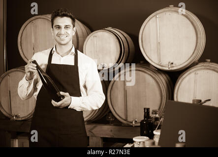 Sorridenti venditore maschio indossando il grembiule in piedi con la bottiglia in mani in cantina Foto Stock