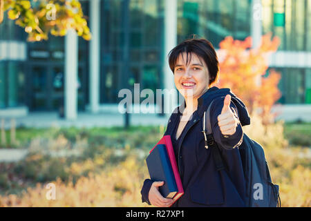 All'aperto autunno Ritratto di giovane donna studente che porta uno zaino e libri che mostra il pollice in alto segno cercando di fotocamera. Foto Stock