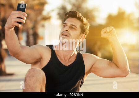 Allegro uomo sportivo che mostra e bicipiti tenendo selfie sulla spiaggia stazione fitness Foto Stock