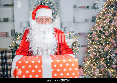 Uomo felice in Sata Claus vestiti si alza e si affaccia sulla fotocamera. Egli detiene il big red box della presente. Non c'è albero di Natale dietro di lui. Foto Stock