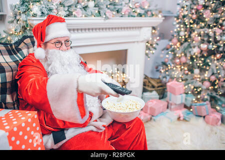 Soddisfatti e felici uomo in Santa Claus costume si siede e utilizza il telecomando. Egli ha una ciotola di popcorn in un altra mano. Ci sono il camino e albero di Natale dietro l'uomo. Foto Stock