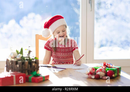 Bambino scrivere la lettera a Babbo Natale. Bambini scrivere Xmas presente la lista dei desideri. Piccola ragazza seduta decorata in soggiorno con una grande finestra su sun Foto Stock