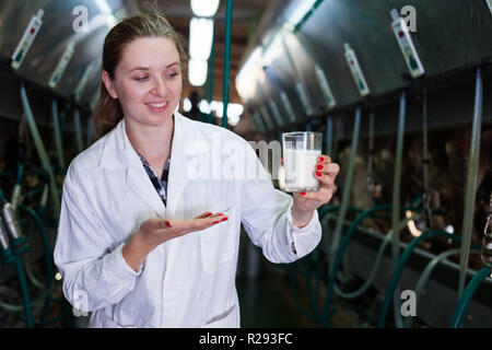 Caseificio femmina ingegnere in tunica bianca in piedi con un bicchiere di latte vicino alla linea di mungitura presso l'azienda Foto Stock