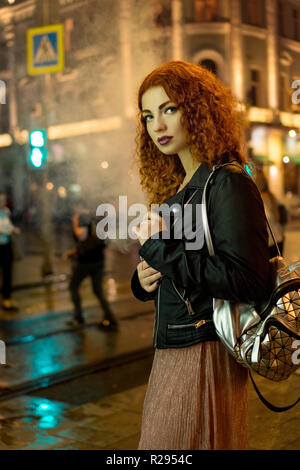 I giovani i Capelli rossi ragazza fuma una sigaretta elettronica. Lonely a piedi per le strade della città di notte. La nicotina sigaretta. Asfalto umido dopo la pioggia. Foto Stock