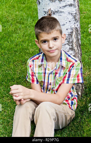 Carino brunette boy undici anni seduto vicino a tree Foto Stock