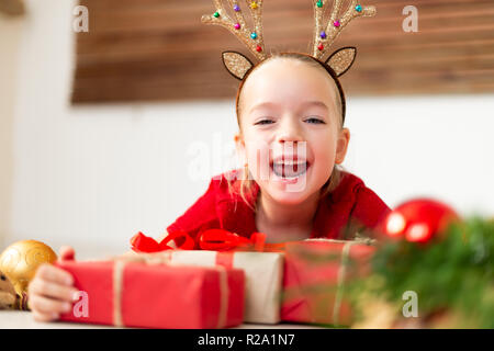 Carino giovane ragazza indossando il costume corna di renne sdraiato sul pavimento, circondato da molti regali di Natale, urla di gioia. Capretto felice a natale Foto Stock