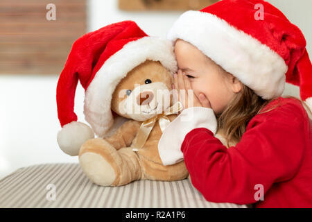 Carino giovane ragazza indossando santa hat sussurra un segreto per il suo orsacchiotto di peluche regalo di Natale giocattolo. Cheeky kid segreti di condivisione con Teddy bear. Foto Stock