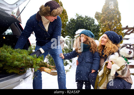 Il padre ha portato ad albero di natale in grande tronco della vettura SUV. Figlia, la madre e il cane incontrare papà felicemente aiutarlo con le vacanze home decorazioni. Famiglia prepara per il nuovo anno insieme. Nevoso inverno all'aperto Foto Stock