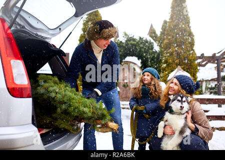 Il padre ha portato ad albero di natale in grande tronco della vettura SUV. Figlia, la madre e il cane incontrare papà felicemente aiutarlo con le vacanze home decorazioni. Famiglia prepara per il nuovo anno insieme. Nevoso inverno all'aperto Foto Stock