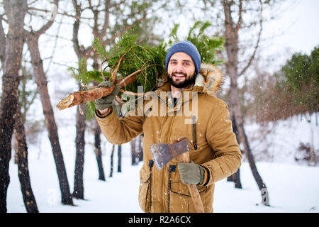 Uomo Barbuto che trasportano appena tagliato all albero di natale nella foresta. Lumberjack contiene ax e abete sulla sua spalla nei boschi. Un comportamento irresponsabile verso la natura, salvare la foresta, mantenere il concetto di verde. Foto Stock