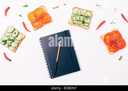 Piano di composizione laici chiuso notepad con coperchio nero vicino al telaio della panini con pomodori e cetrioli, vista dall'alto. Foto Stock
