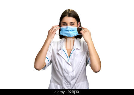 Piuttosto medico mette sulla maschera protettiva. Giovane donna medico mette sulla maschera di medici e guardando la telecamera su sfondo bianco. Foto Stock